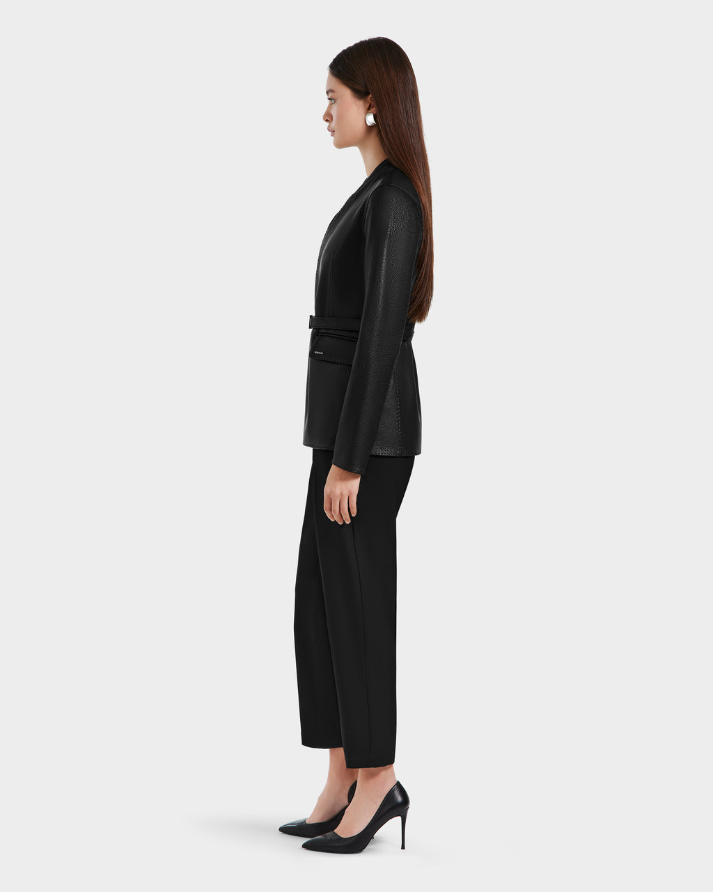 A woman with long brown hair stands in profile, wearing the Rudsak PAMILA - 8125051 Black leather blazer, black pants, and heels—epitomizing minimalist sophistication against a plain light background.
