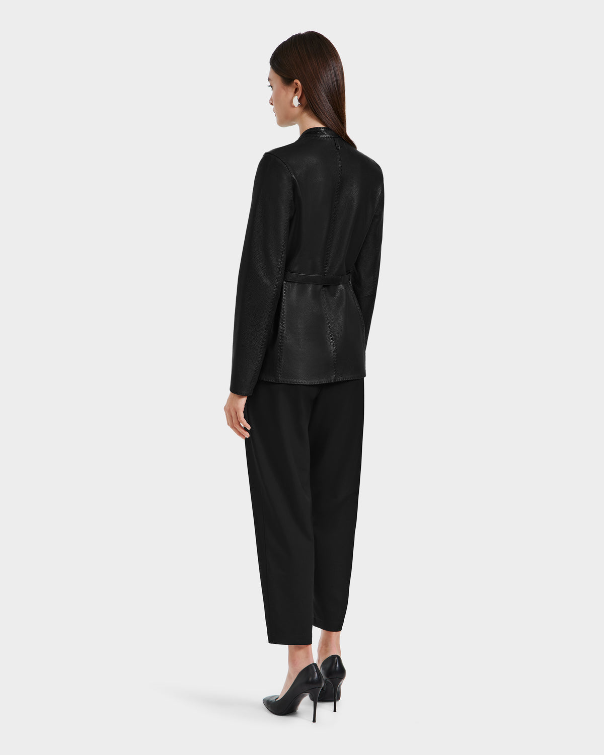 A woman with straight brown hair stands facing away in the Rudsak PAMILA - 8125051 Black womens leather blazer, paired with black pants and high heels, embodying minimalist sophistication against a plain light gray background.