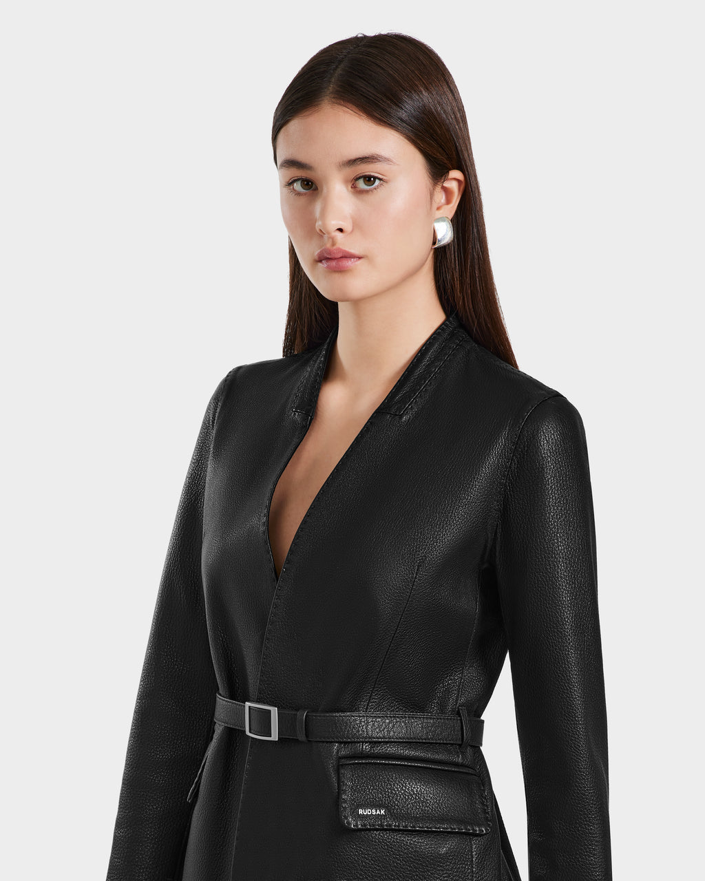 A woman with straight brown hair models the Rudsak PAMILA - 8125051 Black textured leather blazer with a deep V-neck and silver earrings against a light gray background, showcasing minimalist sophistication.