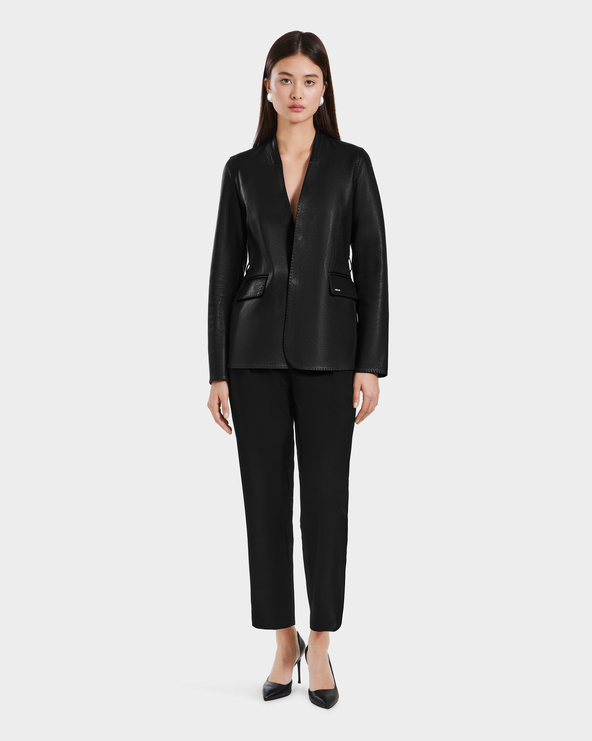 A woman stands facing forward, wearing Rudsaks PAMILA - 8125051 Black textured blazer with black pants and pointed-toe heels against a plain light gray background, embodying minimalist sophistication and versatile style.