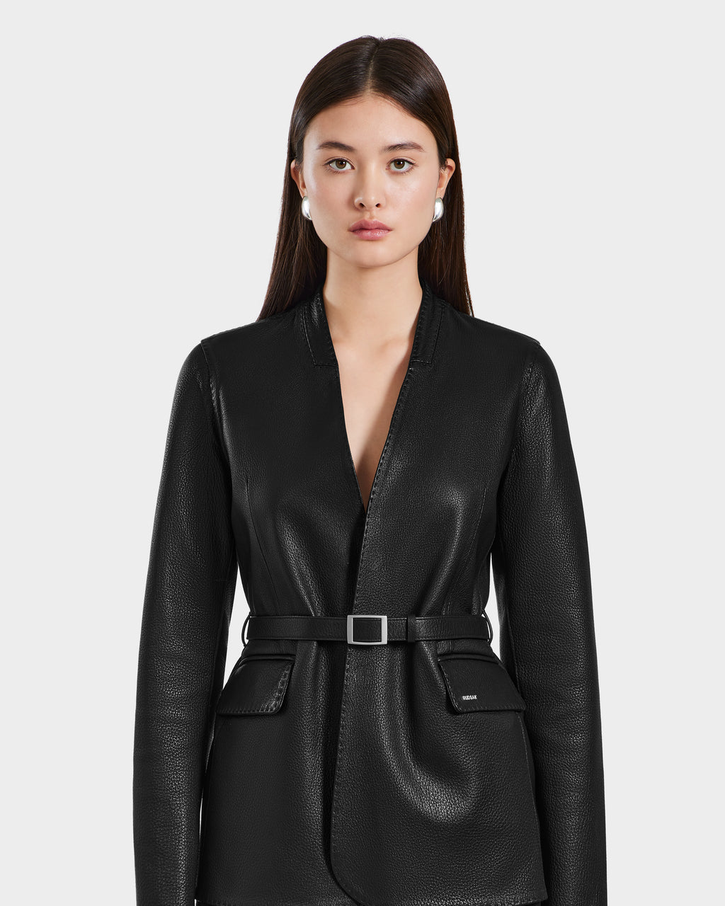 A woman with long, straight brown hair models the Rudsak PAMILA - 8125051 Black textured belted blazer with flap pockets, exuding minimalist sophistication and minimal jewelry, against a plain light background.