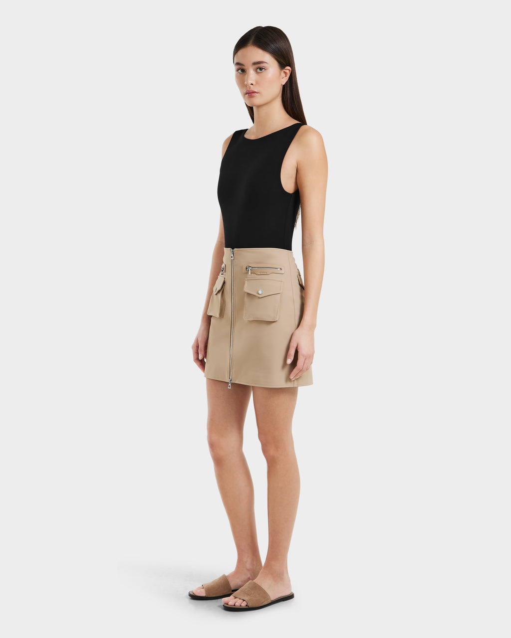 A woman wears a sleeveless black top, Rudsaks PAMINA - 8525025 Almond women’s mini skirt with utility pockets and zippers, and black slide sandals against a plain white background.