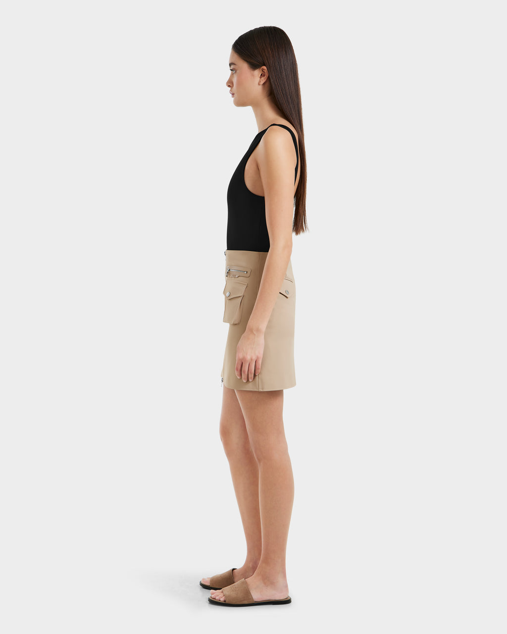 A woman with long straight brown hair stands in profile against a plain background, wearing a black sleeveless top, a beige mini skirt with utility pockets, and Rudsak’s PAMINA - 8525025 Almond brown slide sandals. She appears relaxed.