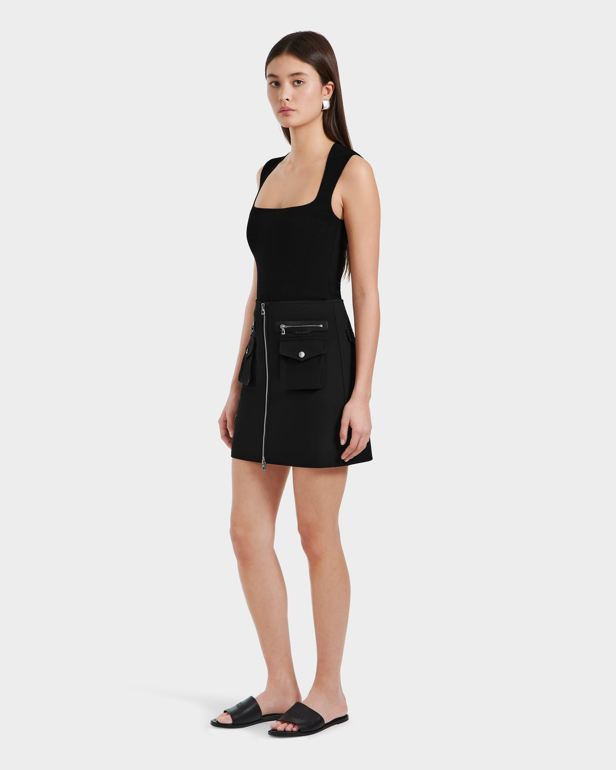 A woman with straight brown hair and a neutral expression wears the Rudsak PAMINA - 8525025 Black sleeveless dress with utility pockets and a zipper, paired with black slide sandals, against a plain background.