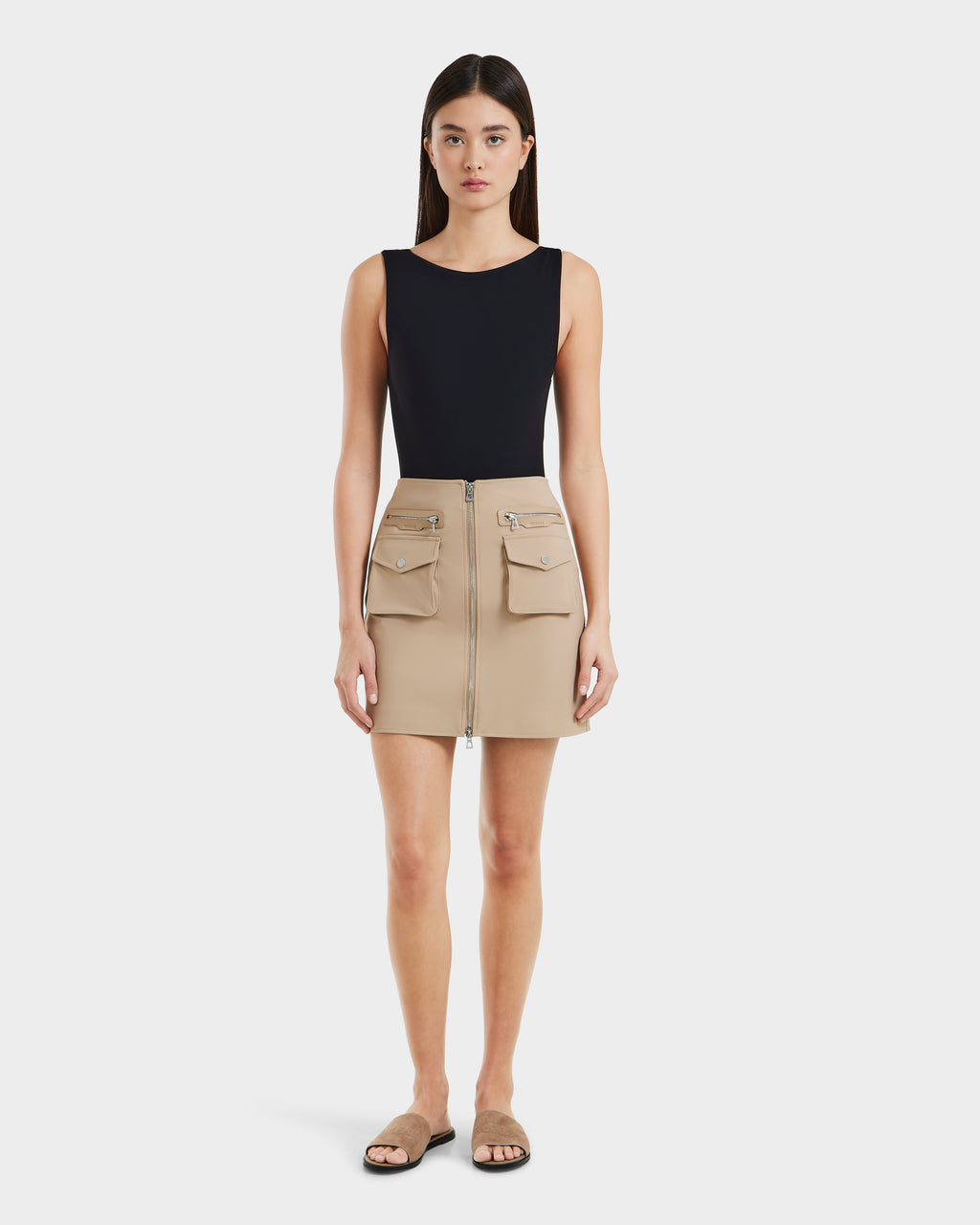 A woman faces forward in a sleeveless black top, beige vegan leather Rudsak PAMINA - 8525025 Almond women’s mini skirt with utility pockets, and beige flat sandals against a plain white background.