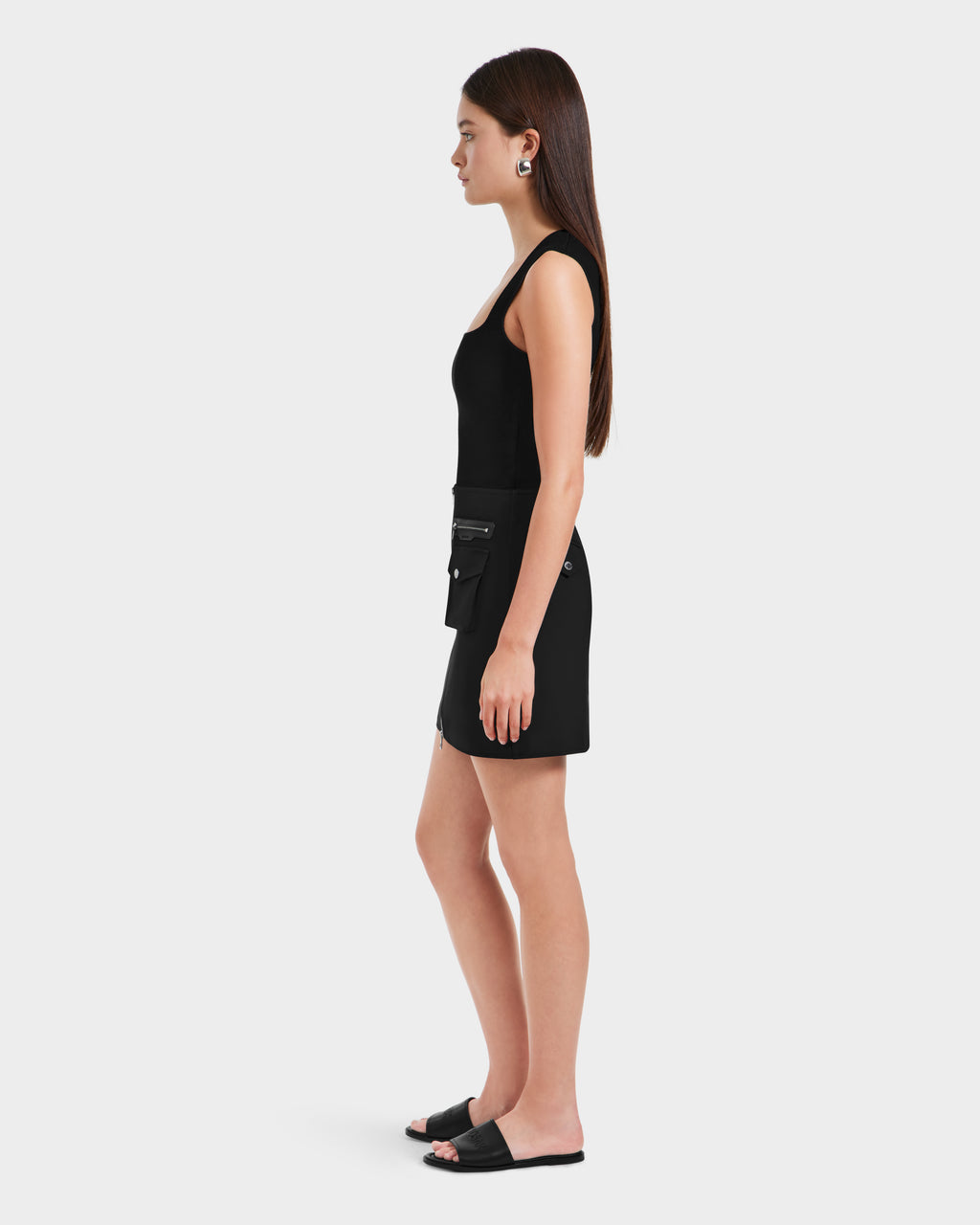 A woman stands in profile against a plain background, wearing the Rudsak PAMINA - 8525025 Black vegan leather womens mini skirt with zipper and utility pocket details, a black sleeveless top, black slide sandals, hoop earrings, and straight hair worn down.
