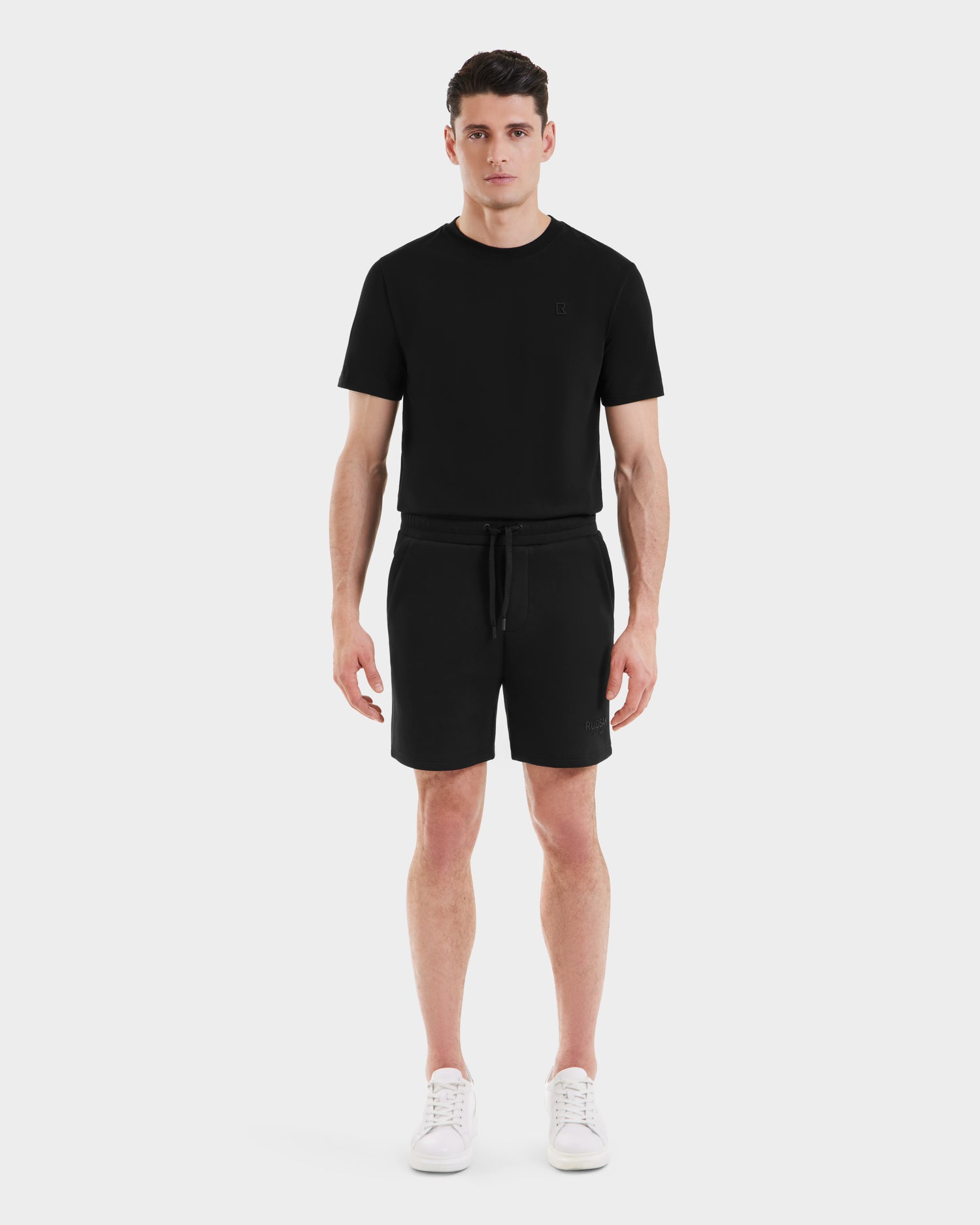 A man stands facing forward against a plain white background, wearing the Rudsak PASCAL - 6524022 Black short-sleeve t-shirt with matching jersey shorts and white sneakers.