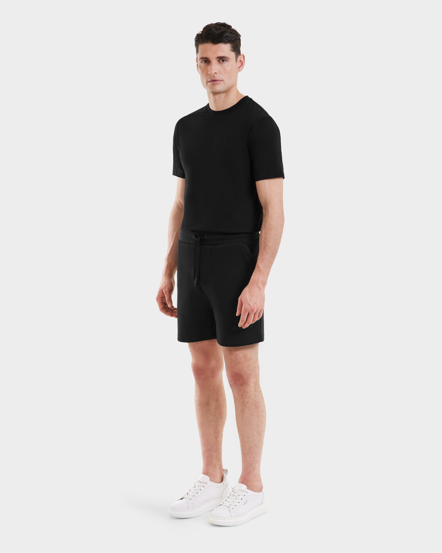 A man stands against a plain white background, wearing the Rudsak PASCAL - 6524022 Black shorts from the BASE Collection, a fitted black T-shirt, and white sneakers. He faces forward with a neutral expression and arms relaxed at his sides.