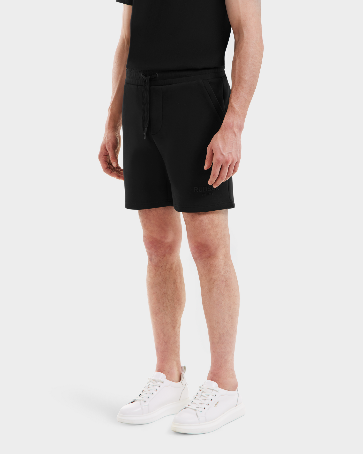 A person wears Rudsak PASCAL - 6524022 Black mens jersey shorts with a stretch waistband, paired with a black t-shirt and white sneakers. The cropped image on a white background shows only the lower torso, legs, and feet.