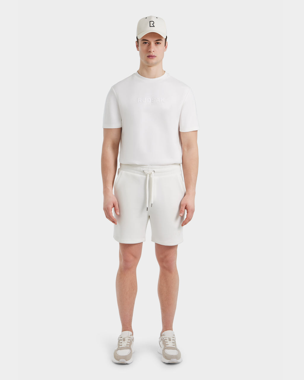 A man stands facing forward against a plain white background wearing the Rudsak PASCAL - 6524022 ICELAND white cap, BASE Collection t-shirt, mens double jersey shorts, and beige sneakers.