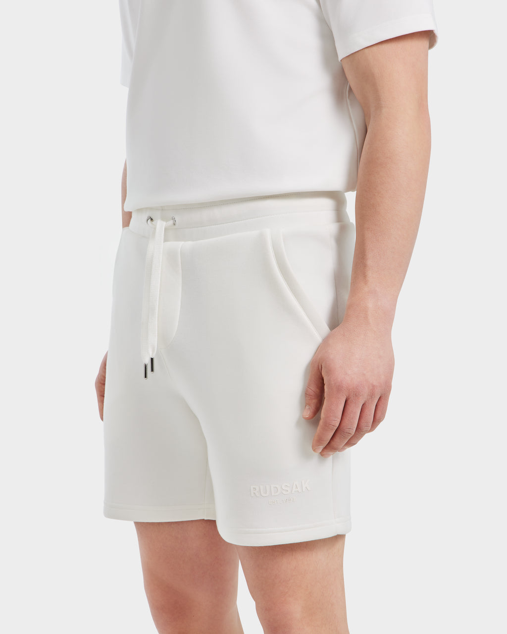 A person wears Rudsaks PASCAL - 6524022 ICELAND mens double jersey shorts with side pockets and RUDSAK embroidery on the left leg, shown in an all-white BASE Collection outfit. Only the lower torso and upper legs are visible.