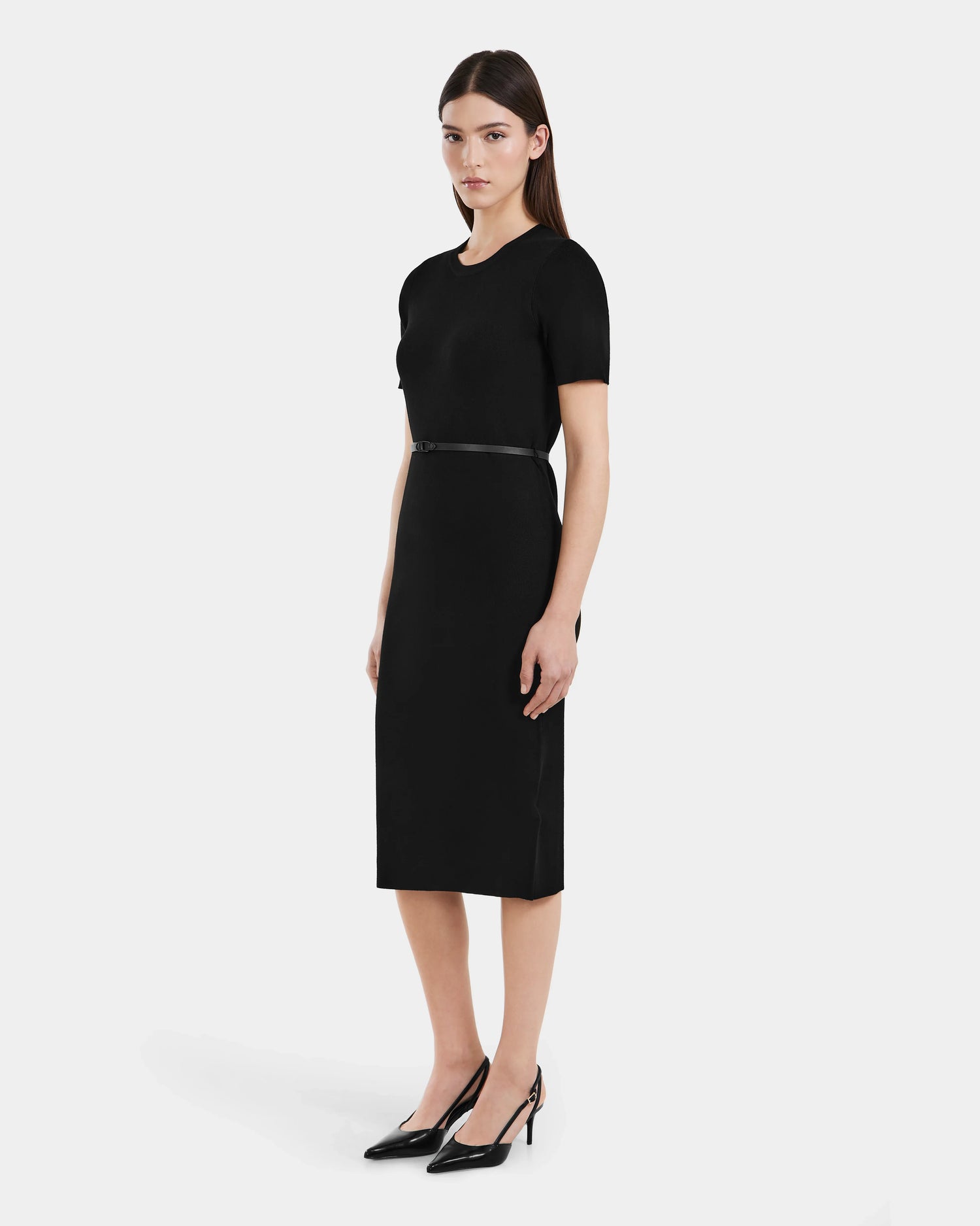 A woman faces slightly to the side against a white background, wearing the PHYLLIS - 8525043 Black fitted knit dress by Rudsak, styled with a vegan leather belt and black heels.
