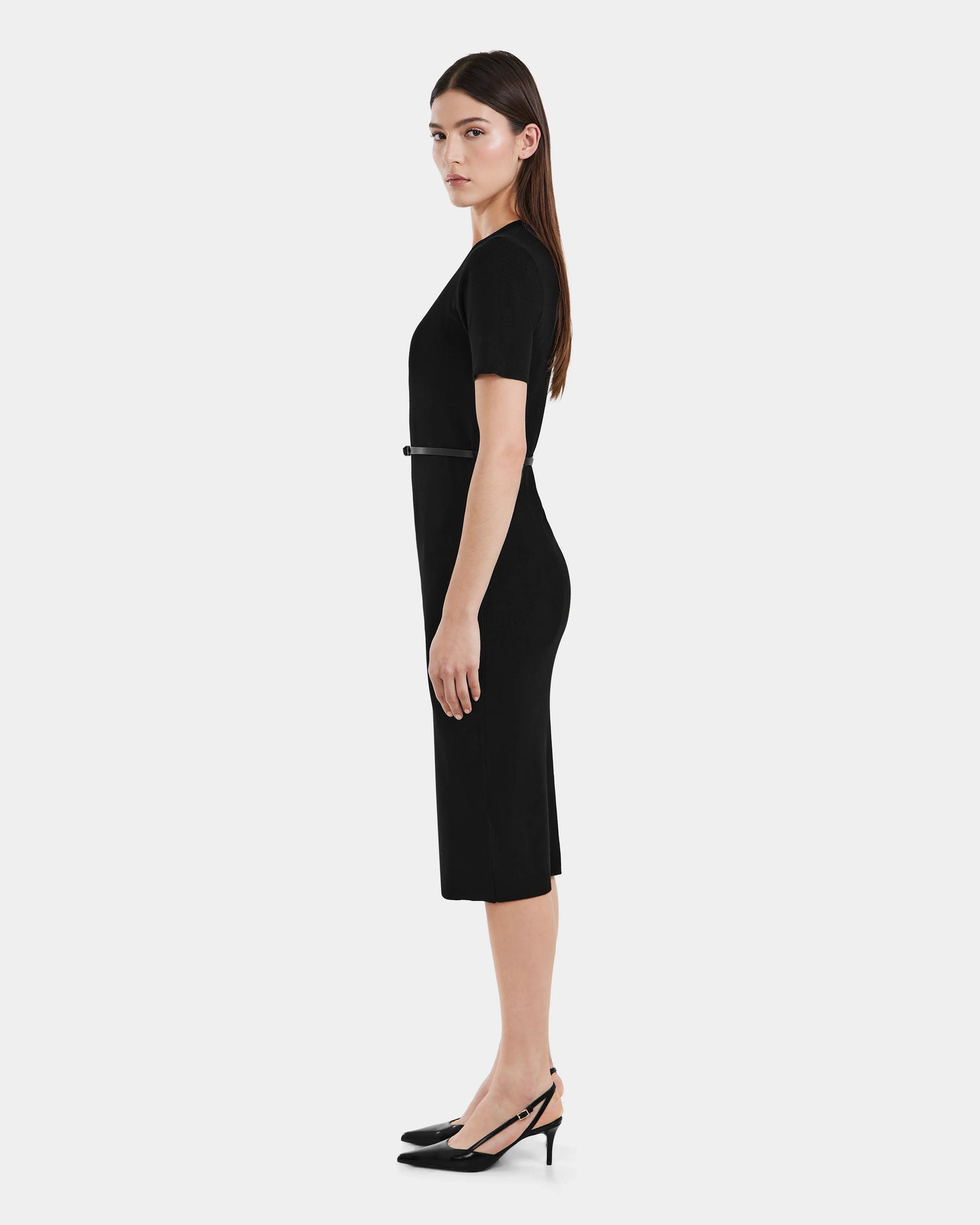 A woman stands in profile against a white background, wearing Rudsaks PHYLLIS - 8525043 Black fitted womens knit dress with short sleeves, a vegan leather belt at the waist, and black slingback heels.