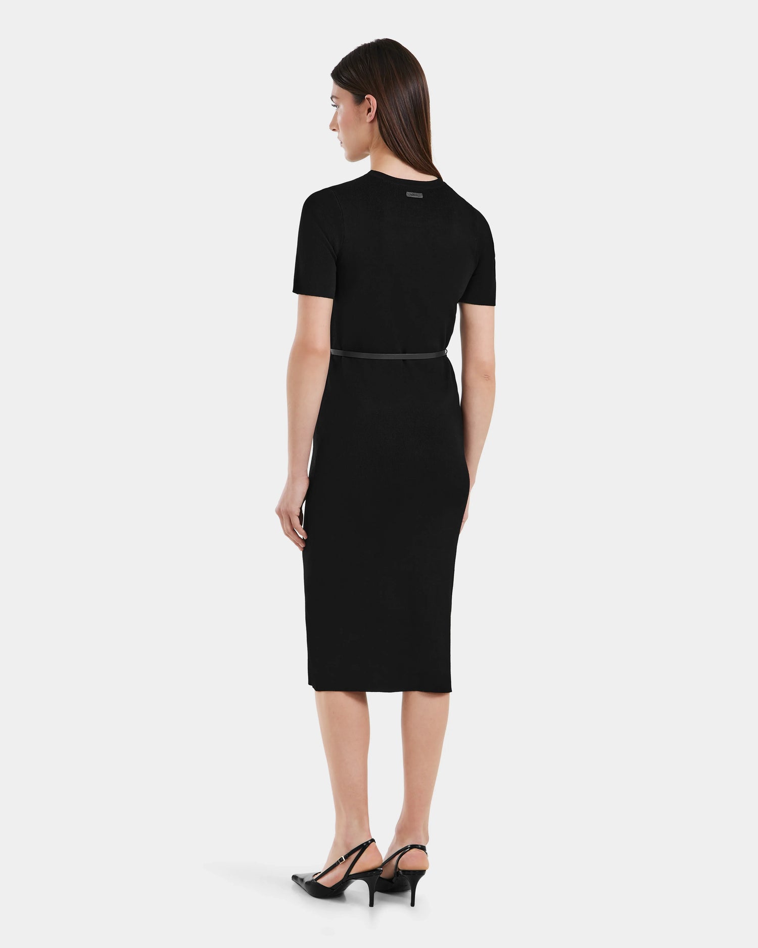 A woman with straight brown hair faces away, wearing the Rudsak PHYLLIS - 8525043 Black fitted women’s knit dress with short sleeves and a thin vegan leather belt, paired with black heeled shoes against a plain white background.