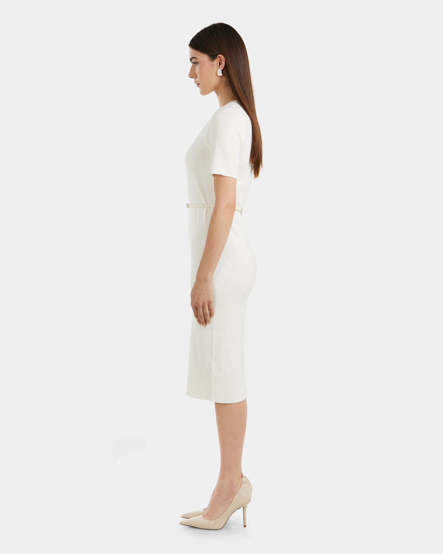A woman with straight brown hair stands in profile wearing the Rudsak PHYLLIS - 8525043 Iceland women’s knit dress with a vegan leather waist belt and beige heels, set against a plain white background.