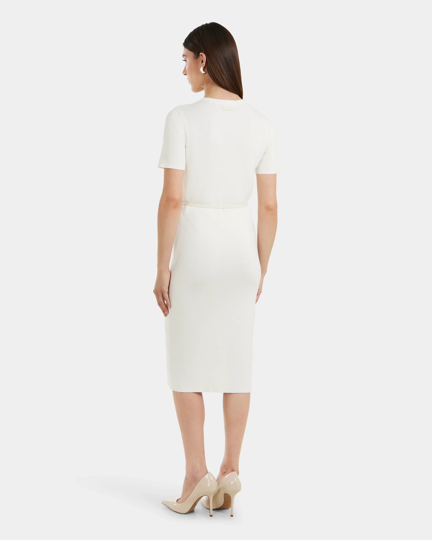 A woman faces away, wearing the Rudsak PHYLLIS - 8525043 Iceland knee-length fitted white knit dress with a vegan leather waist belt and beige heels, set against a plain light background.