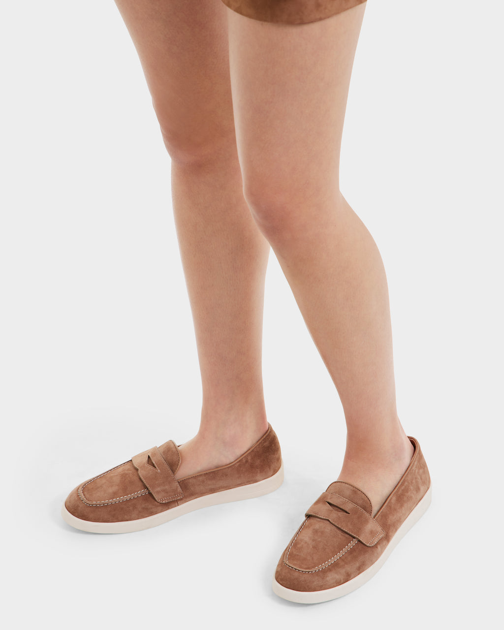 A person’s lower legs and feet are shown wearing Rudsak RABECCA - 8225006 Almond light brown suede slip-on loafers with white soles, highlighting this women’s luxury footwear against a white background.
