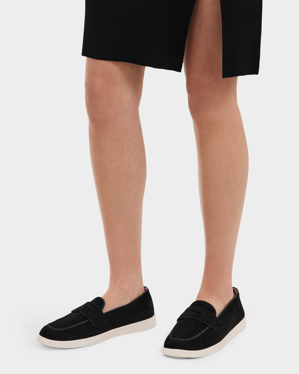 A person stands against a white background wearing a black skirt with a front slit and Rudsak RABECCA - 8225006 Black slip-on loafers, styled as womens luxury shoes with white soles.