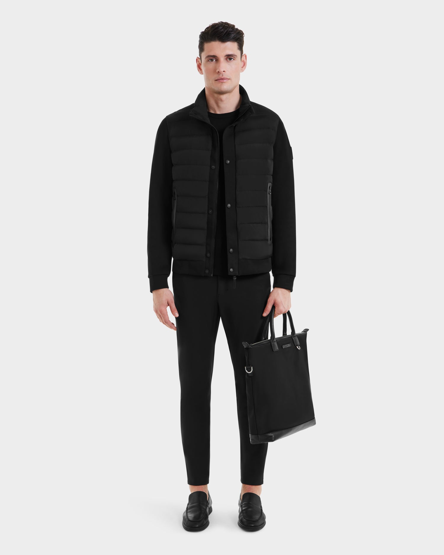A man faces forward wearing the Rudsak RAGAB - 6124577 Black mens hybrid jacket with lightweight down insulation, paired with black pants and shoes. He holds a black tote bag against a light gray background—ideal for transitional weather.