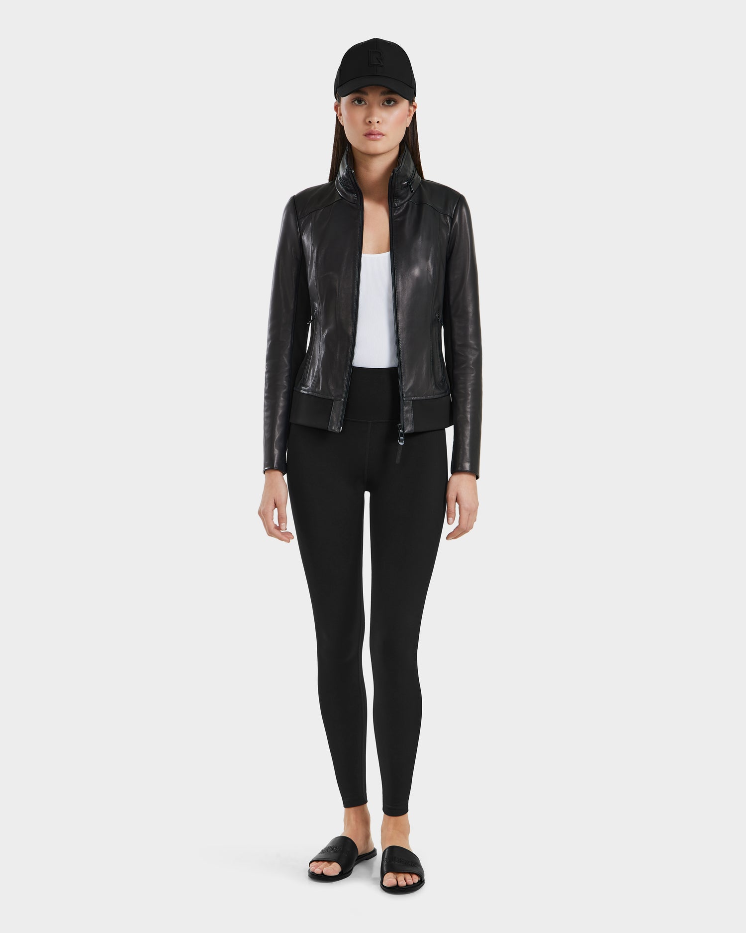 A woman faces forward wearing the Rudsak REBECCA - 8125066 Black lamb leather jacket with a lightweight hideaway hood, white top, black leggings, black slide sandals, and a black baseball cap against a plain light background.