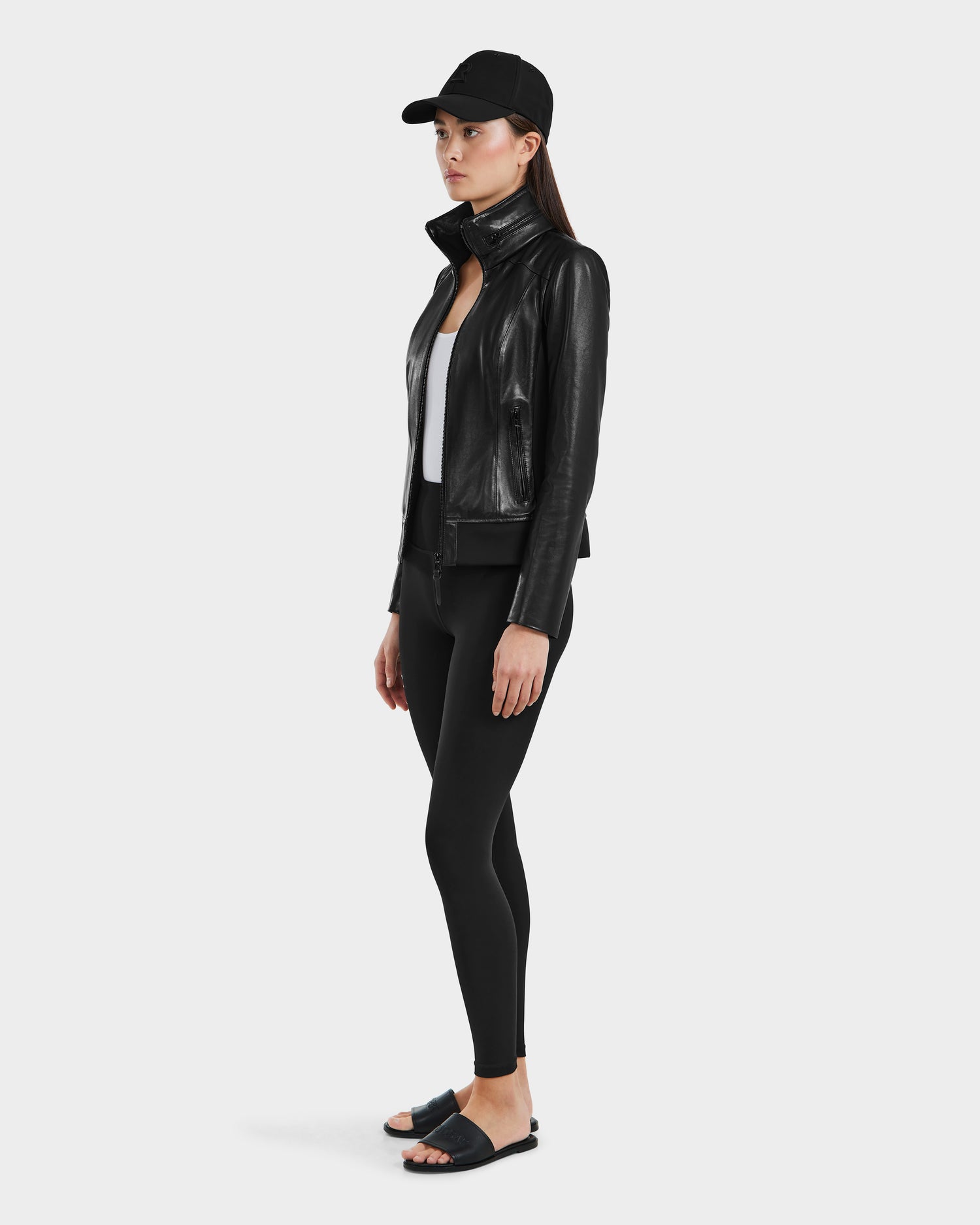 A woman faces left against a plain light background, wearing Rudsak's REBECCA - 8125066 Black lamb leather moto bomber jacket, paired with a black baseball cap, white top, black leggings, and black slide sandals.