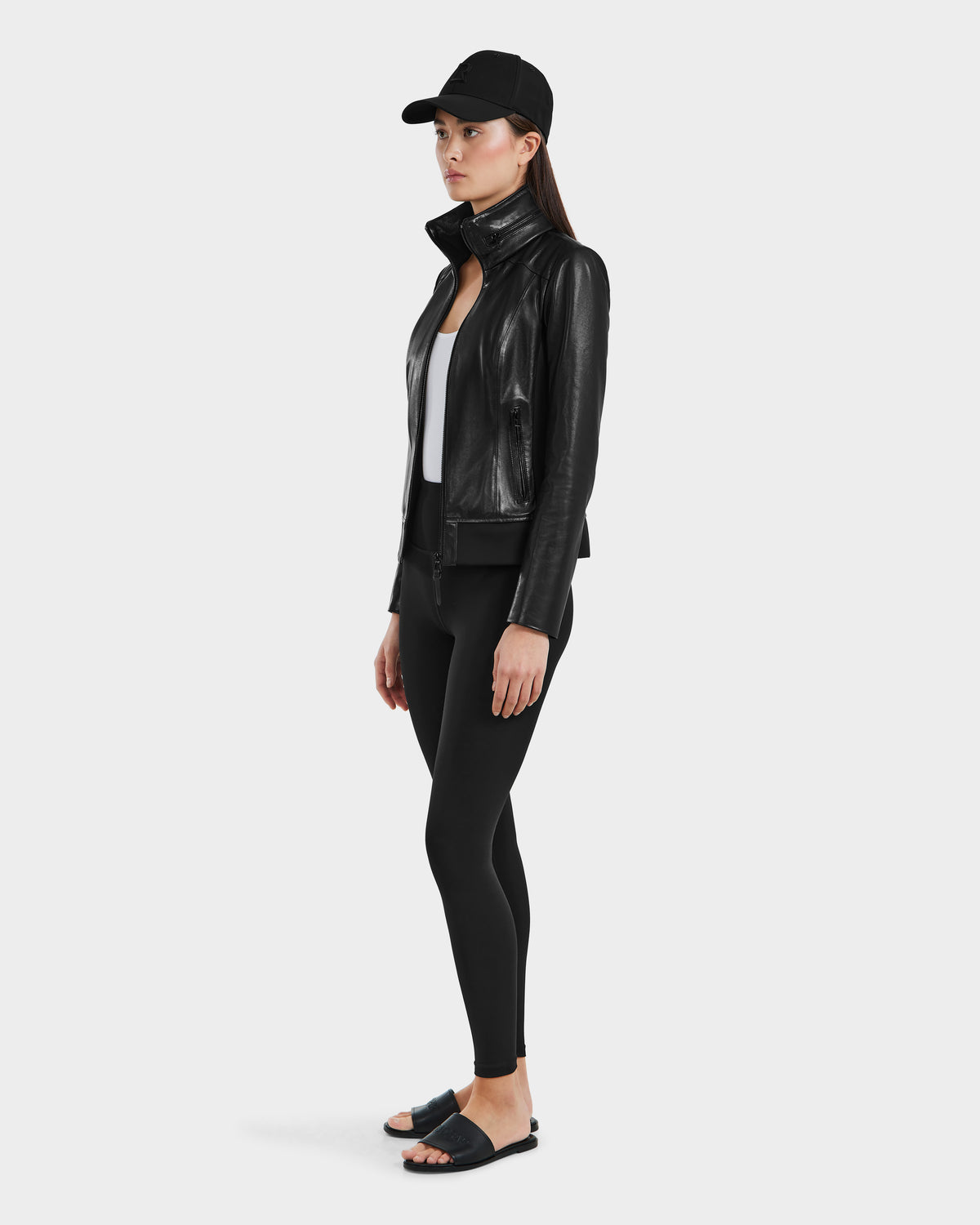A woman faces left against a plain light background, wearing Rudsak's REBECCA - 8125066 Black lamb leather moto bomber jacket, paired with a black baseball cap, white top, black leggings, and black slide sandals.