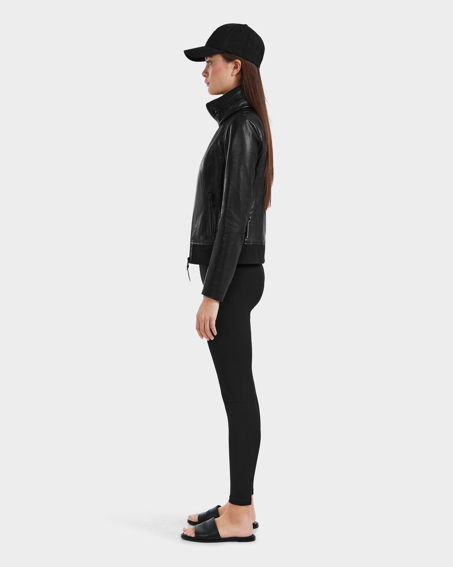 A woman stands in profile wearing the Rudsak REBECCA - 8125066 Black lamb leather jacket with a lightweight hideaway hood, paired with black leggings and slide sandals, set against a plain light background.