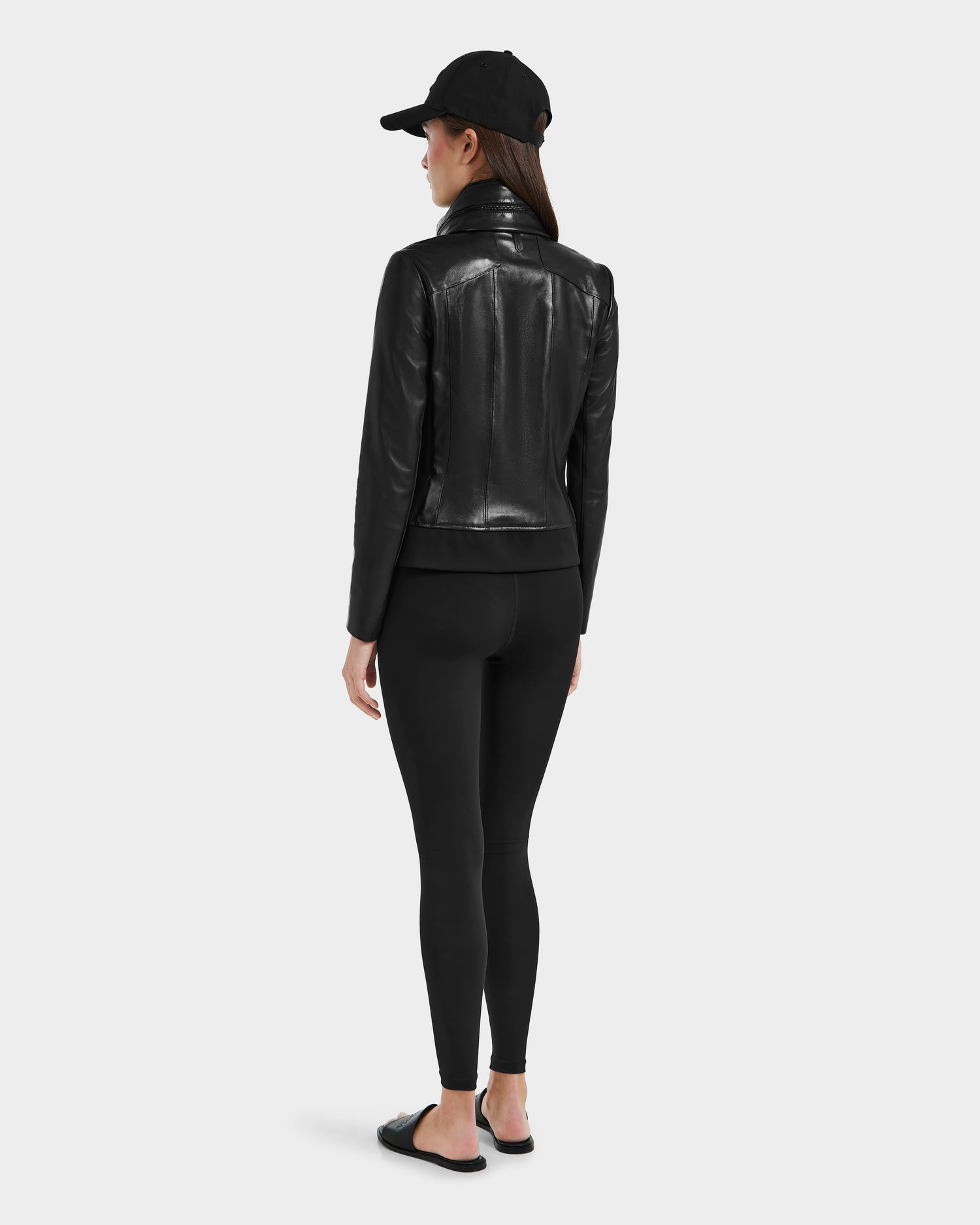 A woman faces away, wearing the Rudsak REBECCA - 8125066 Black moto-inspired bomber lamb leather jacket, paired with black leggings, a black baseball cap, and black slide sandals against a plain light background.