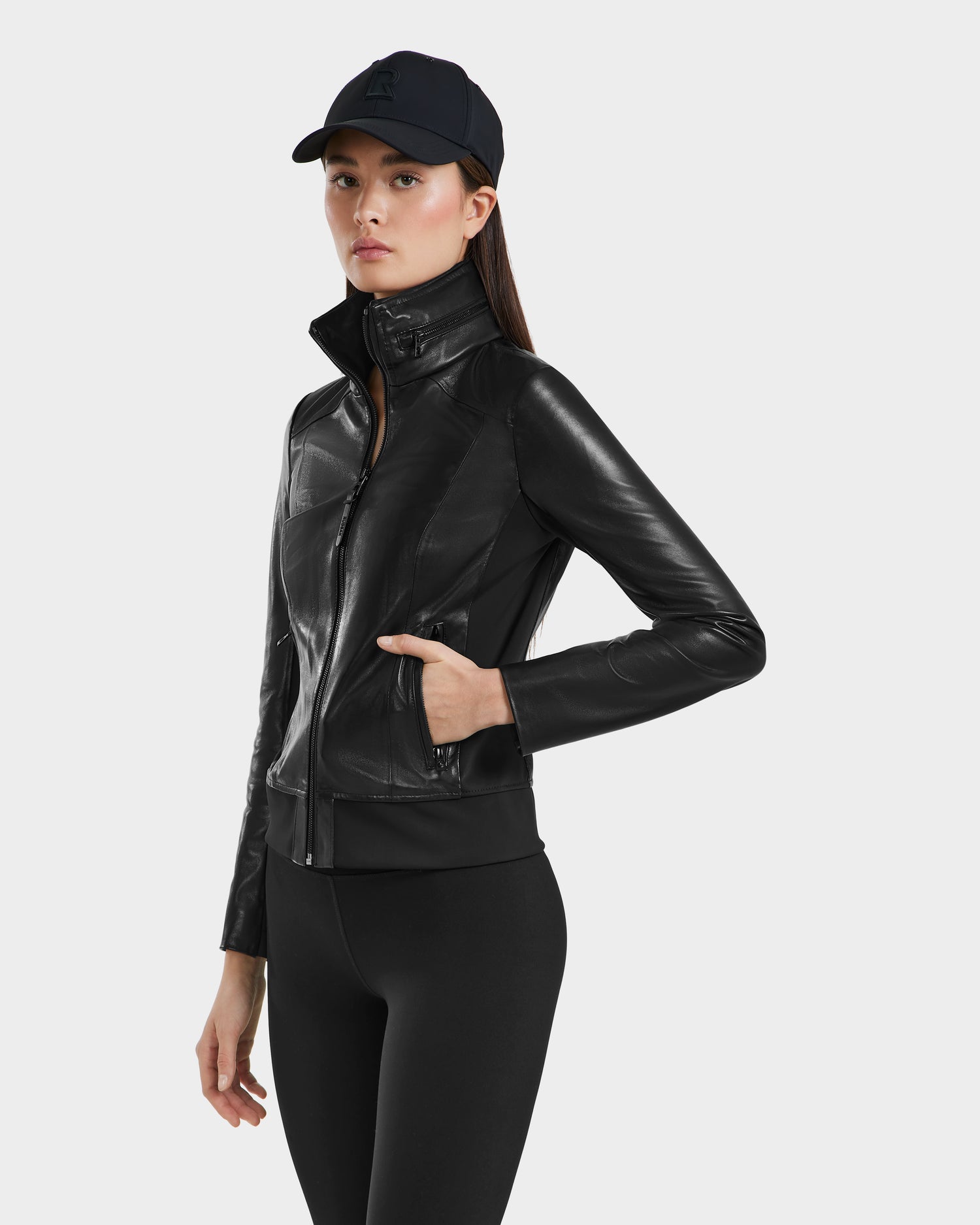 A woman wearing the Rudsak REBECCA - 8125066 Black moto-inspired lamb leather bomber jacket, paired with black leggings and a black baseball cap, stands with one hand in her pocket against a plain light background.