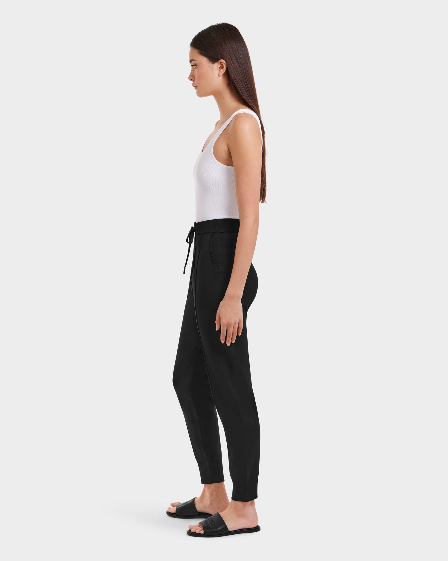 A woman in profile wears a white sleeveless top, high-rise loungewear jogger pants, and Rudsak REBEKA - L525004 Black slide sandals, posed against a plain white background.