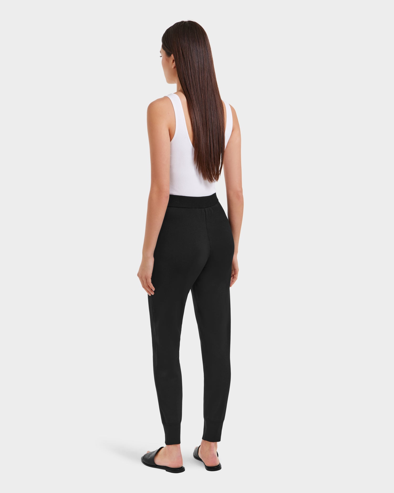 A woman stands facing away against a plain light background, wearing a white sleeveless top and Rudsak REBEKA - L525004 Black high-rise jogger pants, paired with black open-toe slide sandals.