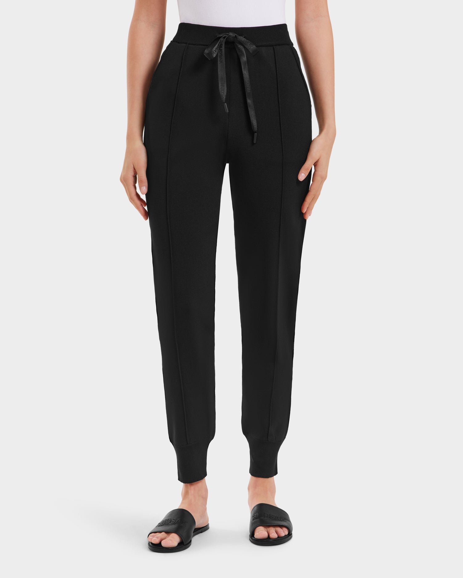 A person wears Rudsak REBEKA - L525004 Black high-rise jogger pants with a drawstring waist, vertical seams, and ribbed cuffs, paired with black slides and a white top, shown from waist down against a plain background.