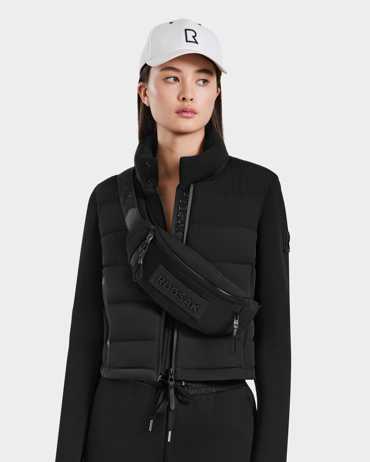 A woman wears a white baseball cap, black puffer vest, black long-sleeve top, and black pants. She carries the Rudsak RILO - 9323008 BLACK crossbody belt bag with waterproof zippers against a plain white background.