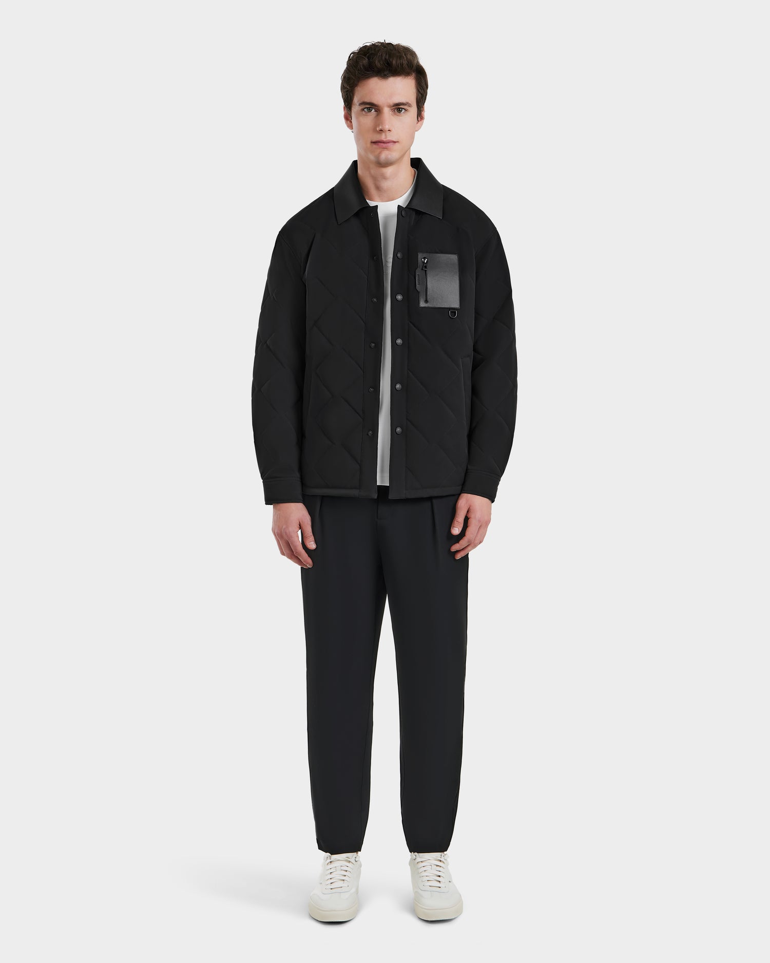 A modern man stands facing forward, wearing the Rudsak RYAN - 6125023 Black quilted light jacket with eco-friendly insulation, black pants, a white shirt underneath, and white sneakers against a plain white background.