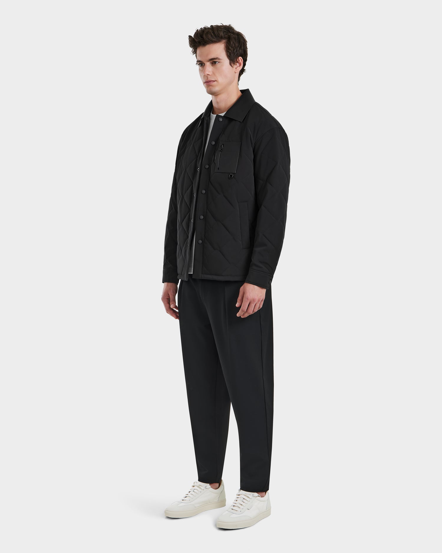 A modern man stands against a white background wearing the Rudsak RYAN - 6125023 Black quilted jacket with eco-friendly insulation, black pants, a gray shirt, and white sneakers. He faces slightly to the side with a neutral expression.