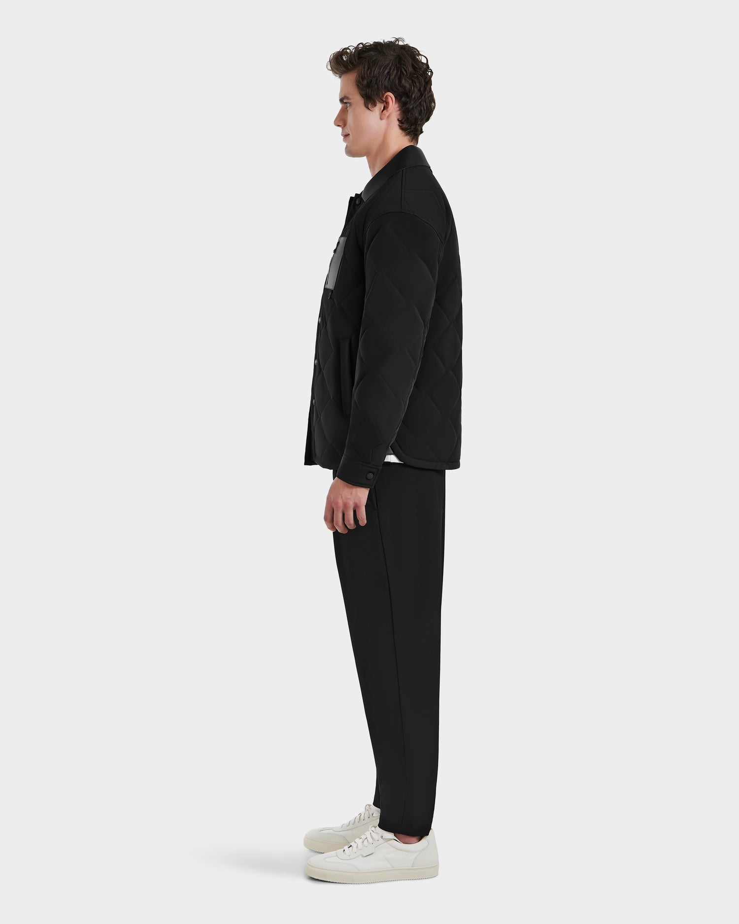 A modern man stands in profile against a plain background, wearing the Rudsak RYAN - 6125023 Black light jacket with eco-friendly insulation, paired with black pants and white sneakers.