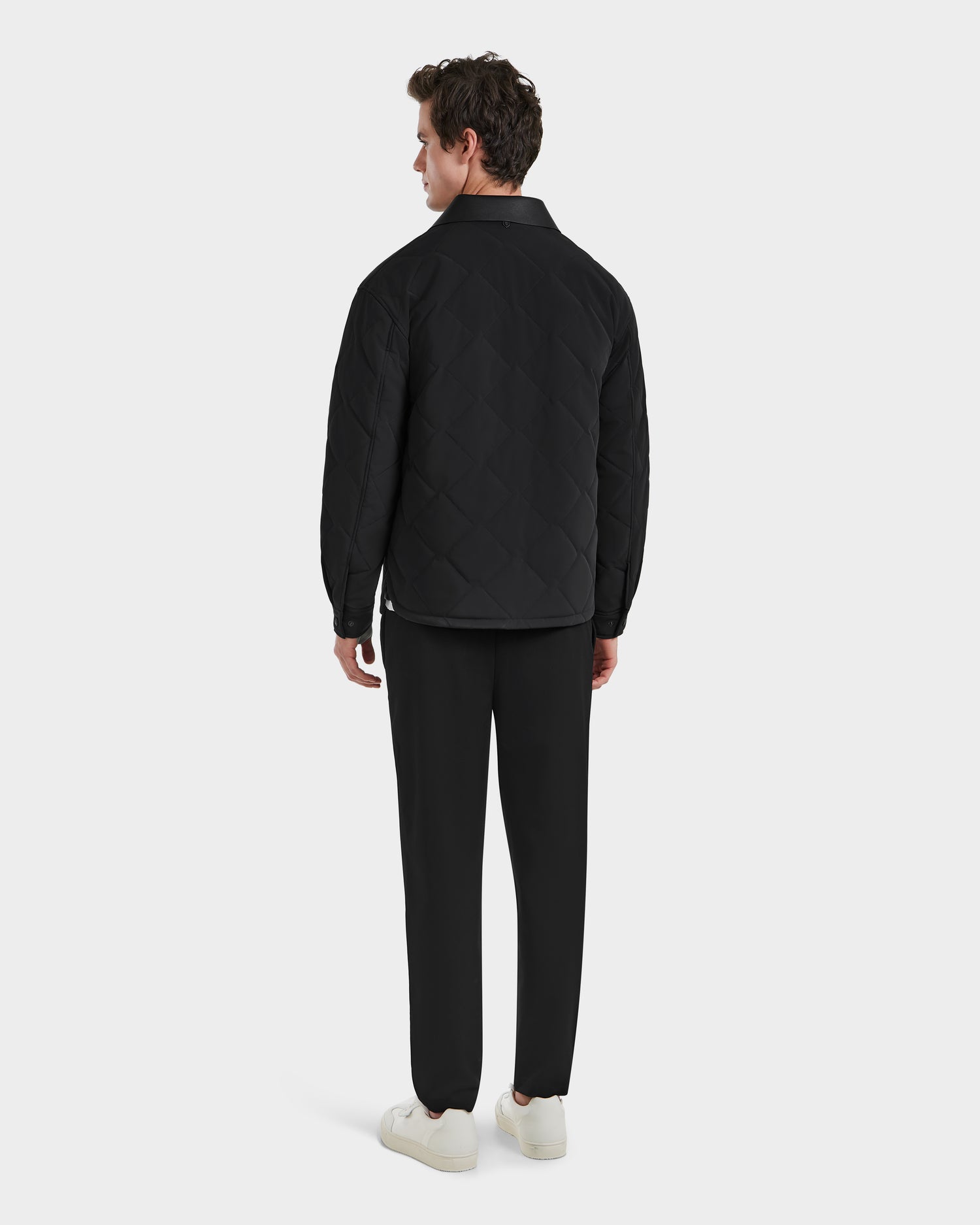 A modern man faces away, wearing the Rudsak RYAN - 6125023 Black quilted jacket featuring eco-friendly insulation, paired with black pants and white sneakers against a plain light background.