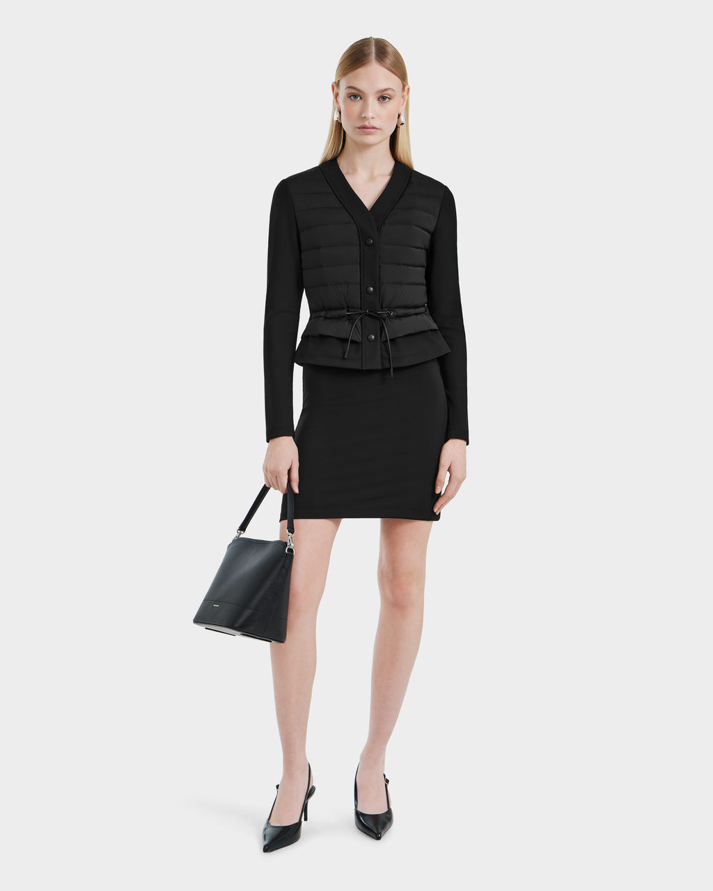 A woman stands against a white background, wearing the Rudsak SAMANTHA - 8125043 Black women’s hybrid cardigan with padded vest and drawstring waist, styled with a fitted black skirt, black heels, and a black handbag.