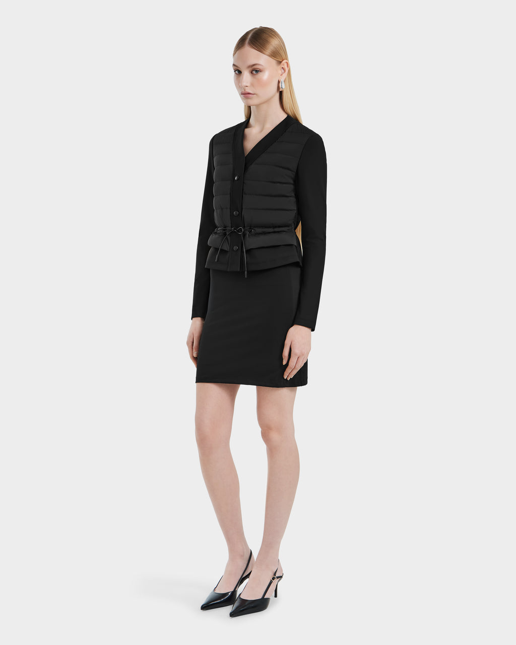 A woman with straight blonde hair wears the Rudsak SAMANTHA - 8125043 Black women’s hybrid cardigan, styled as a quilted down vest with a drawstring waist, over a black long-sleeve dress and black pointed-toe heels against a white background.
