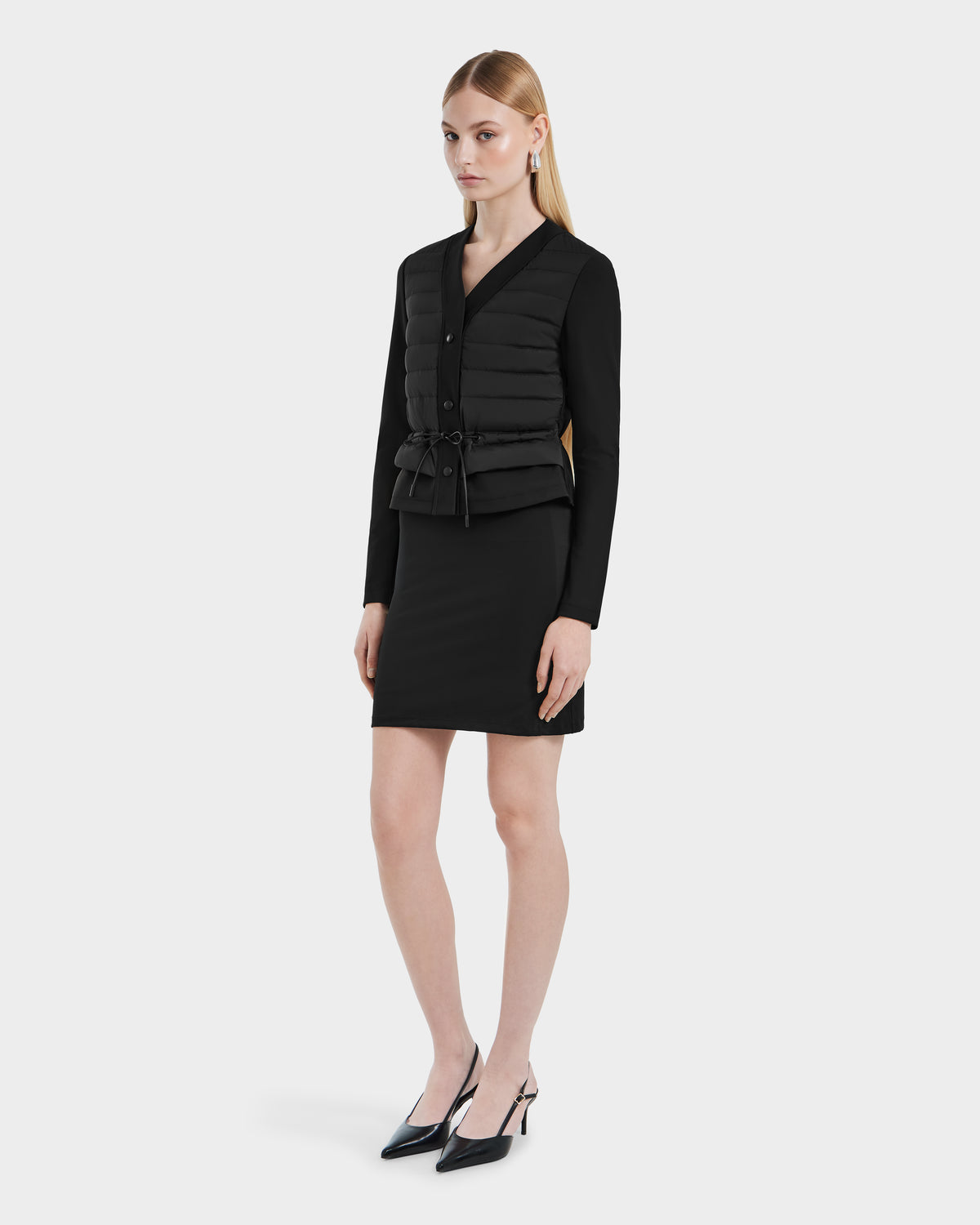 A woman with straight blonde hair wears the Rudsak SAMANTHA - 8125043 Black women’s hybrid cardigan, styled as a quilted down vest with a drawstring waist, over a black long-sleeve dress and black pointed-toe heels against a white background.