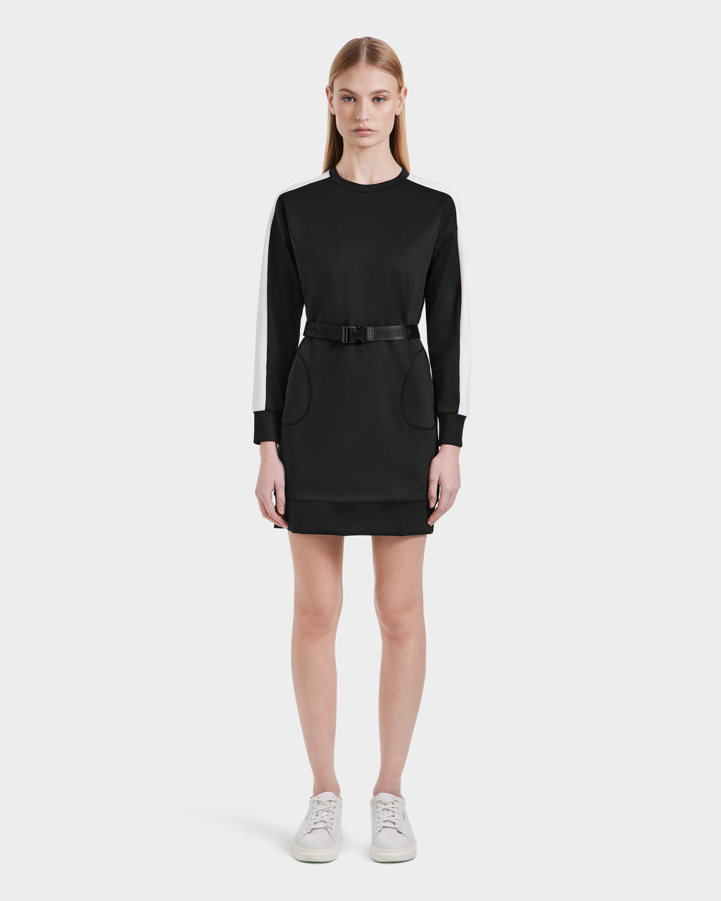 A woman with straight light brown hair and a neutral expression models the Rudsak SETA - 8525019 Black dress, a relaxed-fit black long-sleeve style with white striped sleeves, matching belt, and white sneakers against a plain white background.