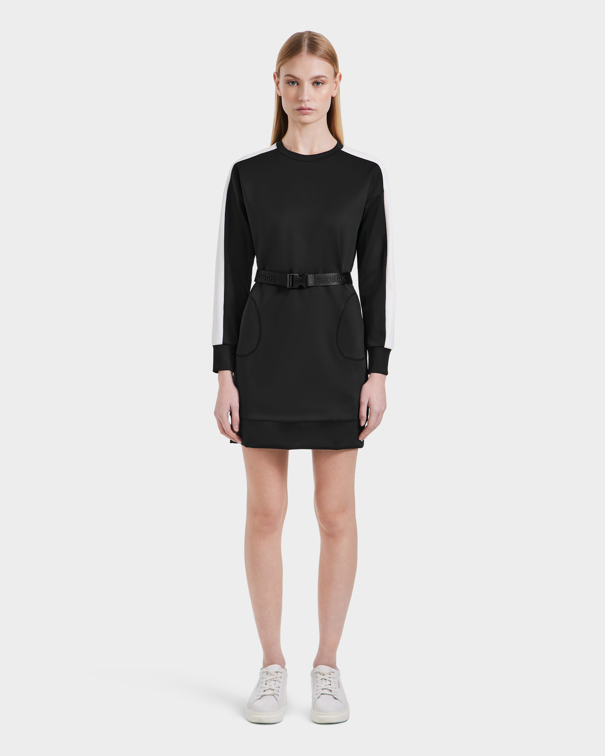 A woman with straight light brown hair and a neutral expression models the Rudsak SETA - 8525019 Black dress, a relaxed-fit black long-sleeve style with white striped sleeves, matching belt, and white sneakers against a plain white background.
