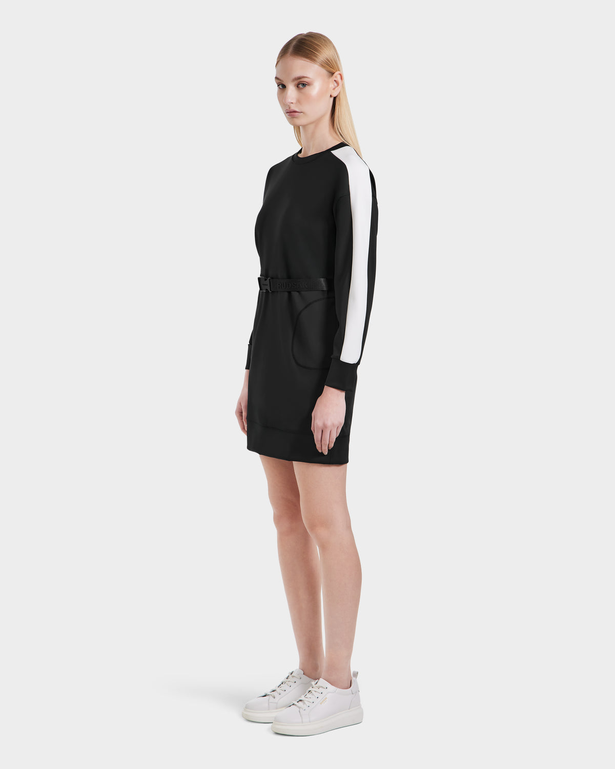 A woman models the Rudsak SETA - 8525019 Black dress—a women’s relaxed-fit, long-sleeve design with white striped sleeves and a belted waist—styled with white sneakers against a simple light background.