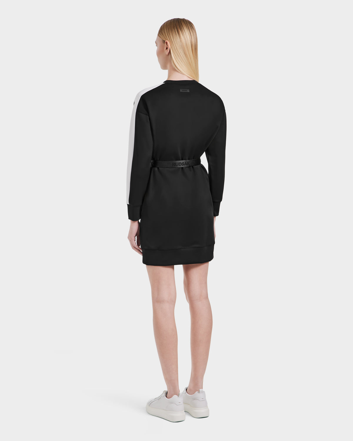 A person with straight blond hair, facing away, wears the Rudsak SETA - 8525019 Black womens relaxed-fit dress with a white sleeve stripe, black belt, and white sneakers—highlighting versatile styling against a plain light backdrop.
