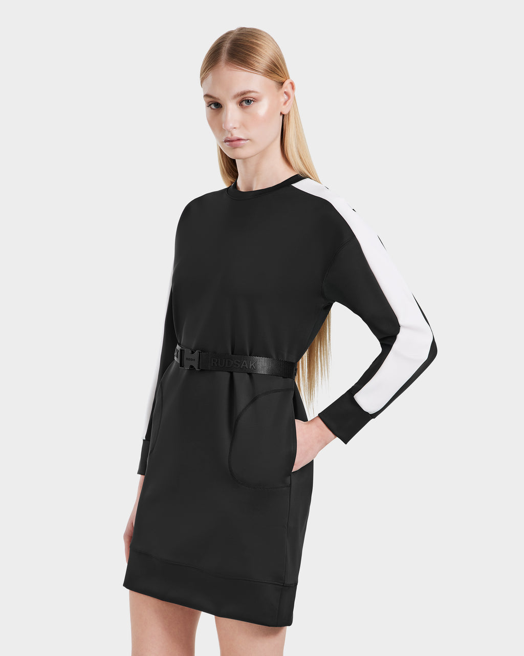 A woman with long, straight blonde hair models the Rudsak SETA - 8525019 Black dress, a relaxed-fit women’s style with white striped long sleeves and a black belt, creating a minimalist silhouette against a plain light background.