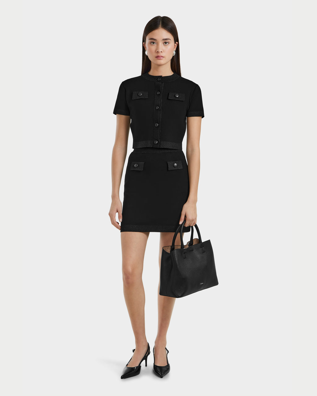 A woman models the Rudsak SIDNEY - 8525074 Black dress with front pockets, styled with black heels and a handbag. Her straight brown hair and neutral expression enhance the contemporary knitwear look against a plain background.