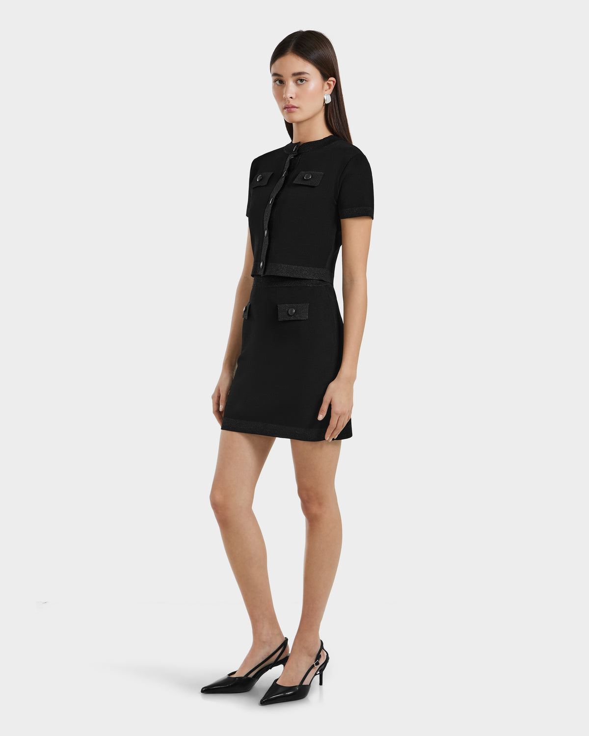 A woman wears the Rudsak SIDNEY - 8525074 Black short-sleeve dress with front pockets and a belted waist, paired with black pointed-toe heels. She has straight brown hair and stands against a plain white background, embodying modern knitwear style.