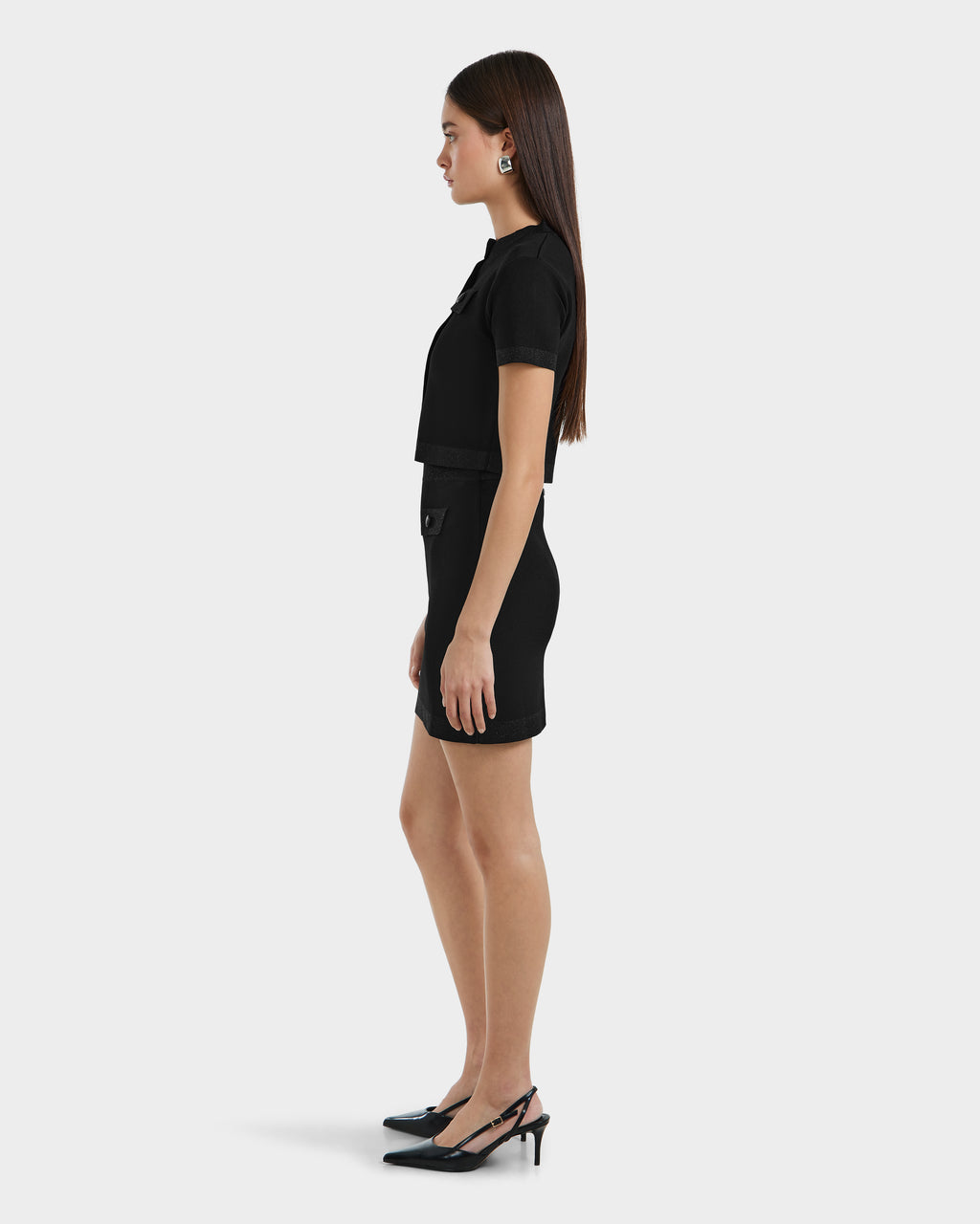 A woman stands in profile wearing the Rudsak SIDNEY - 8525074 Black fitted knit top and matching skirt, paired with black slingback heels and large hoop earrings. Her straight brown hair contrasts with the plain light background.