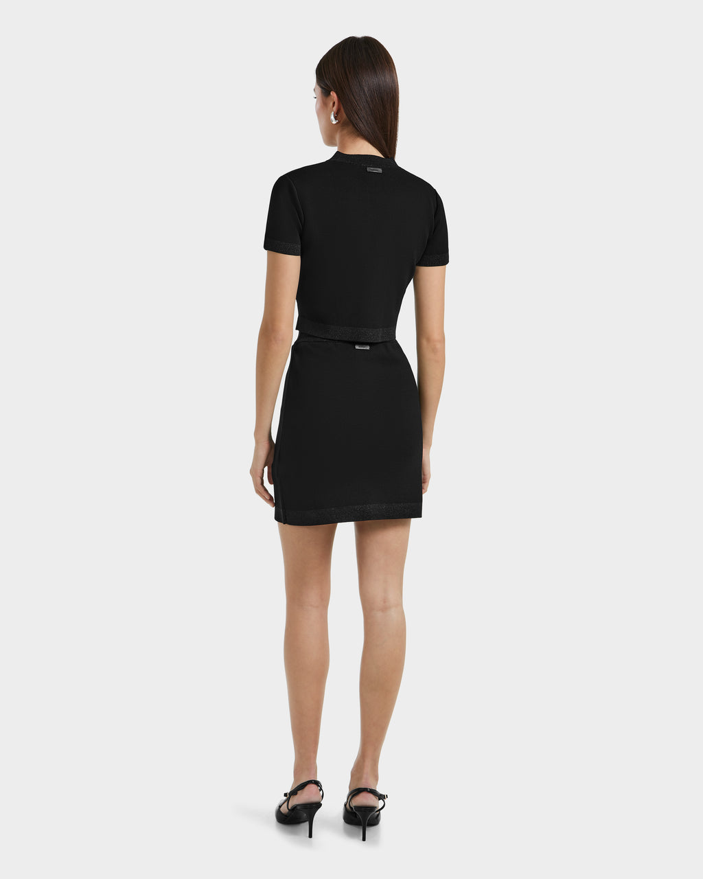 A woman with long brown hair wears the Rudsak SIDNEY - 8525074 Black fitted cropped sweater with metallic trim over a black dress and heeled sandals, standing against a light background for a modern look.