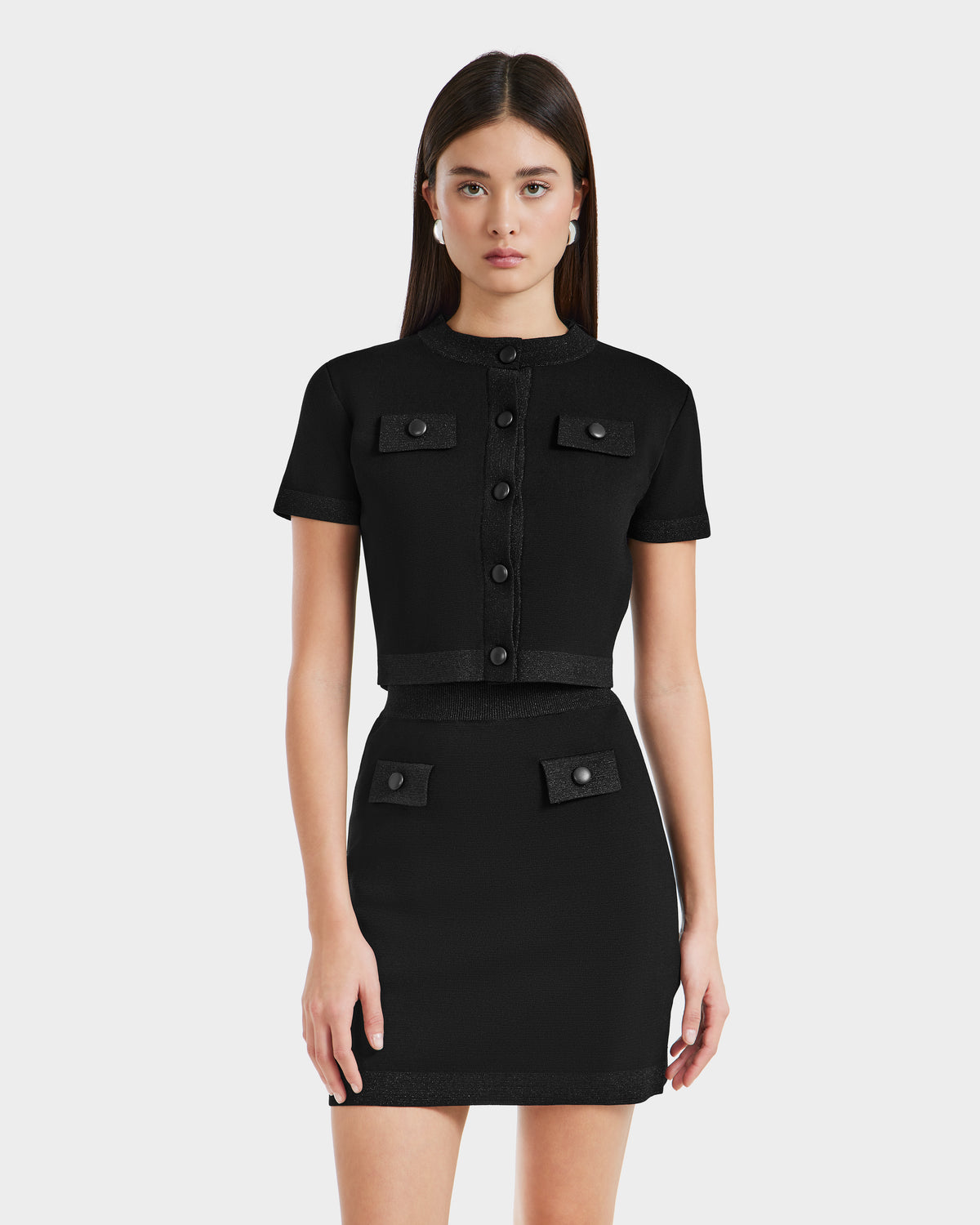 A young woman with straight brown hair wears the Rudsak SIDNEY - 8525074 Black fitted cropped sweater over a short-sleeve black button-up crop top and matching mini skirt with decorative pockets, standing against a plain light background.